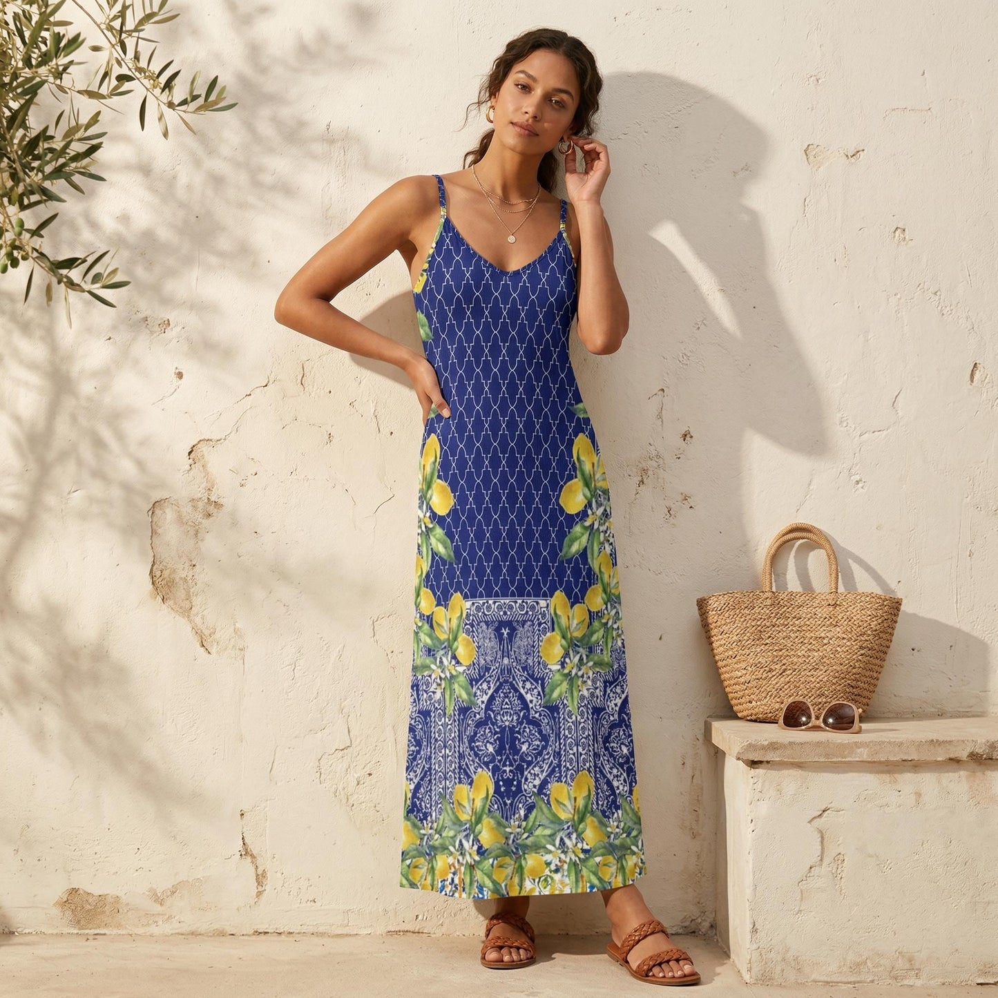 May include: A blue maxi dress with a white geometric pattern and lemon print. The dress has spaghetti straps and a V-neck. A straw tote bag and sunglasses sit on a stone bench.
