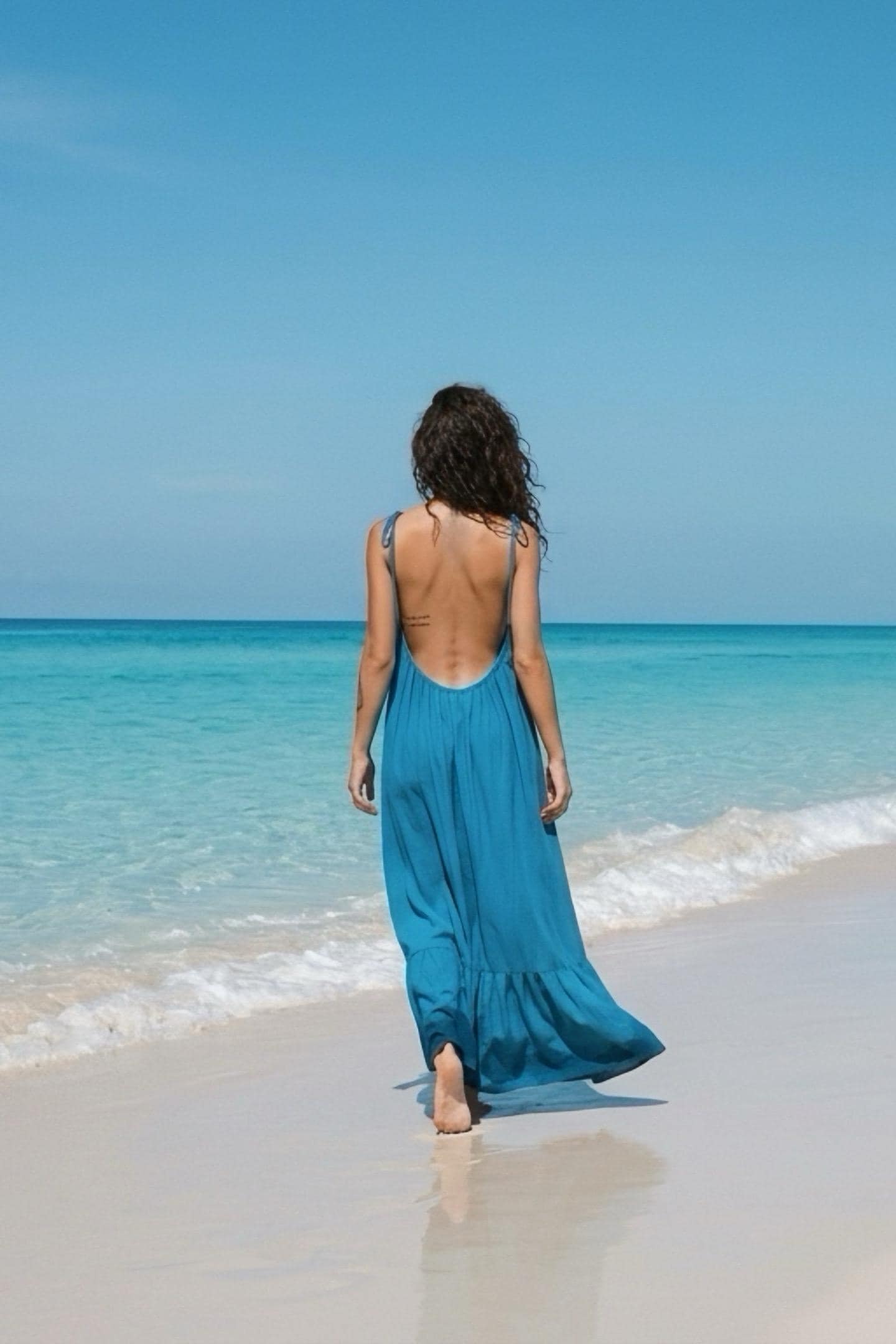Woman wearing a blue turquoise open back maxi dress with tie shoulders and a loose flowy silhouette on a beach, styled as resort wear for vacation.
