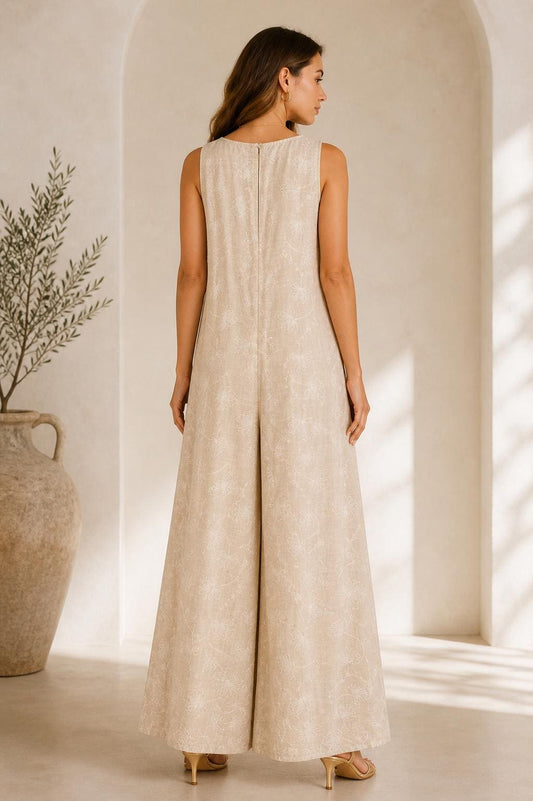 May include: A sleeveless, wide-leg jumpsuit in a neutral beige hue. The garment features a subtle, textured pattern and a zipper closure at the back. The model is wearing gold-colored heels. The background includes a large vase and a plant.