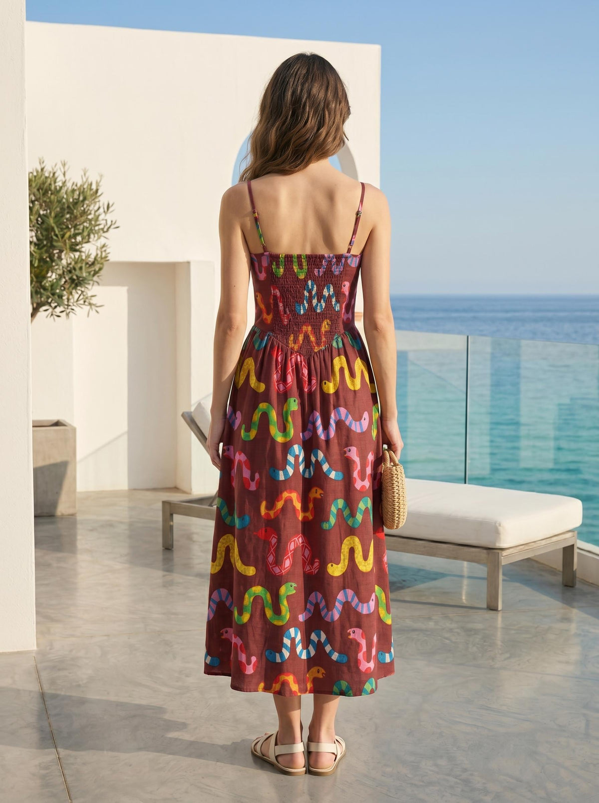 May include: A brown midi dress with colorful snake patterns. The dress features thin straps, a smocked bodice, and a flowing skirt. The model is wearing white sandals and holding a small woven bag. The background includes a patio and ocean view.