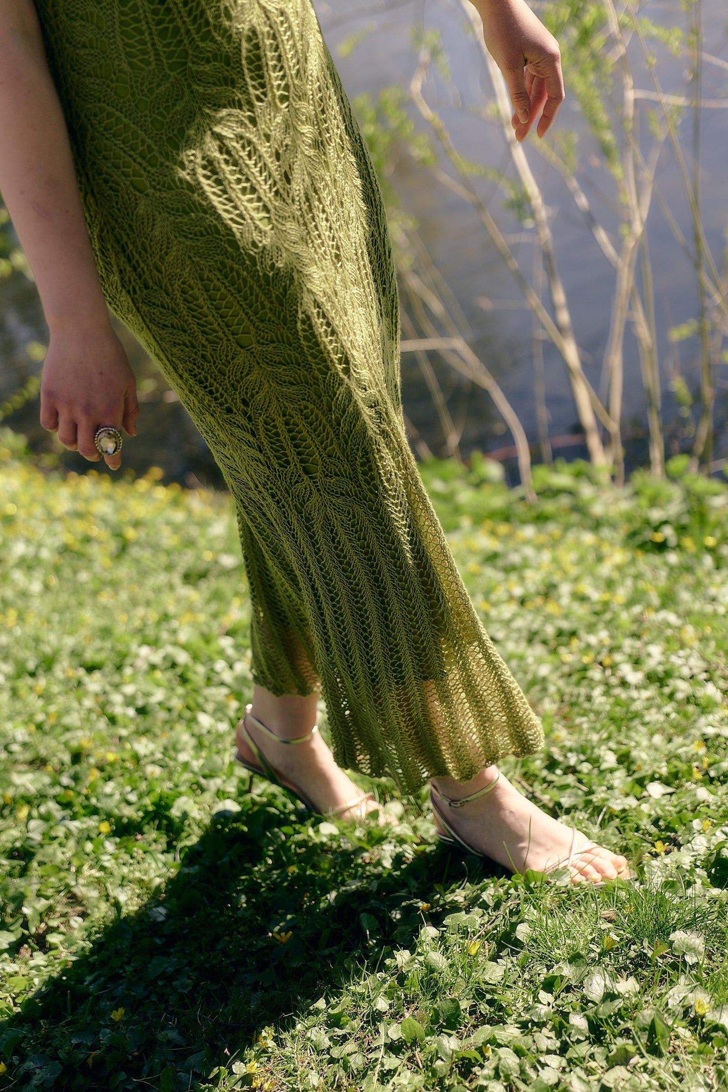 May include: A long, olive green knitted dress with a detailed leaf pattern. The dress is worn with gold sandals. The person is standing on a grassy area with a shadow cast on the grass. The dress appears to be handmade.