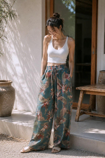 May include: A woman wearing a white ribbed tank top and wide-leg pants. The pants are teal with a floral pattern in shades of red, pink, and beige. She is standing outside near a wooden bench and a large ceramic pot.