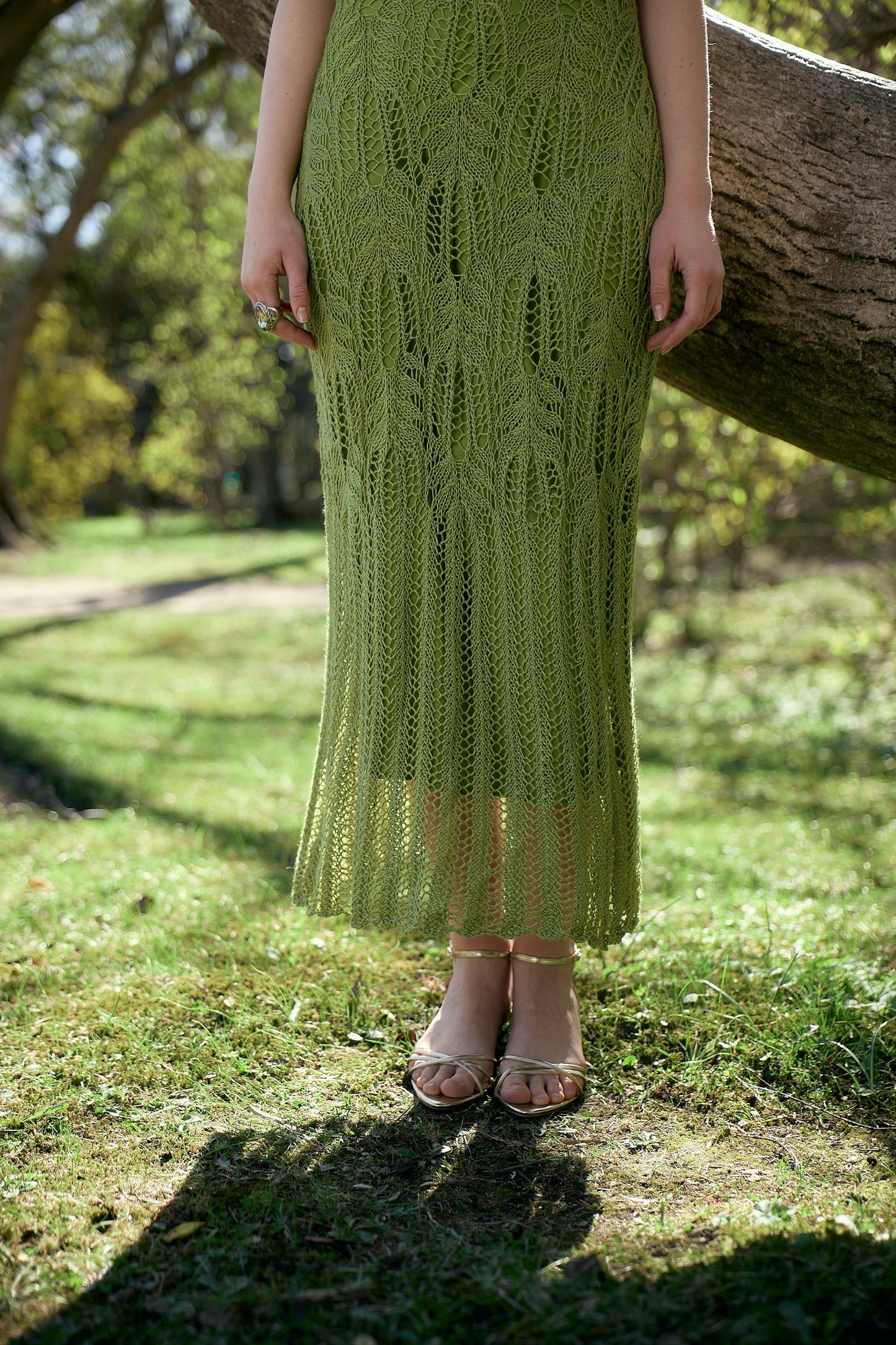 May include: A light green, open-weave knit dress with a leaf-like pattern. The dress falls to the ankles and is paired with gold sandals. The background is a grassy area with a tree.