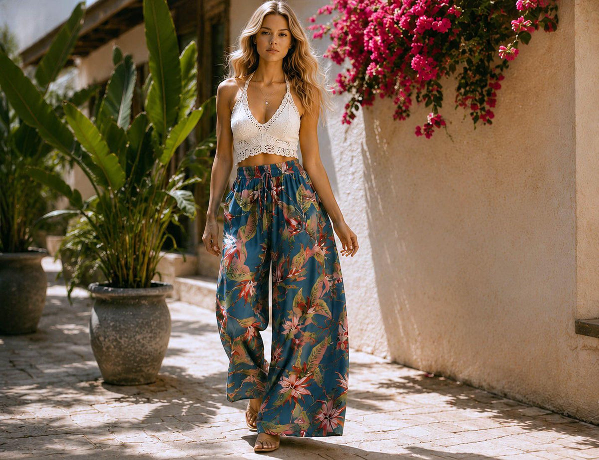 May include: A woman wearing a white crochet crop top and wide-leg pants with a blue floral pattern. The pants have a drawstring waist. The background includes large potted plants and a wall with pink flowers.
