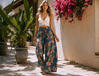 May include: A woman wearing a white crochet crop top and wide-leg pants with a blue floral pattern. The pants have a drawstring waist. The background includes large potted plants and a wall with pink flowers.