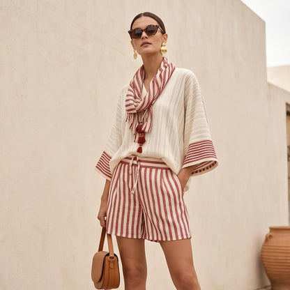 May include: A cream-colored top with wide sleeves and vertical stripes, paired with matching cream and red vertical striped shorts featuring a drawstring waist. A coordinating striped scarf with red tassels hangs around the neck, and a tan handbag is held.