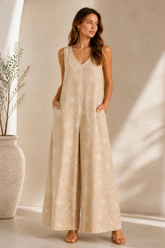 May include: A beige sleeveless jumpsuit with a wide leg and a delicate white pattern. It has a V-neck and pockets. The model is wearing gold sandals. The outfit is suitable for casual or semi-formal occasions. The jumpsuit is made of a light fabric.