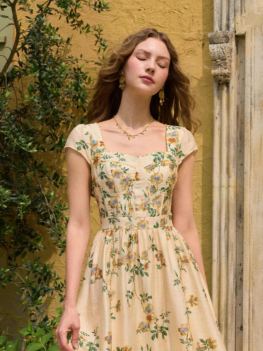 May include: A cream-colored midi dress featuring a square neckline, short cap sleeves, and a fitted bodice with vertical seams. The garment is decorated with an all-over floral print in shades of yellow, blue, and green with leafy vine details.
