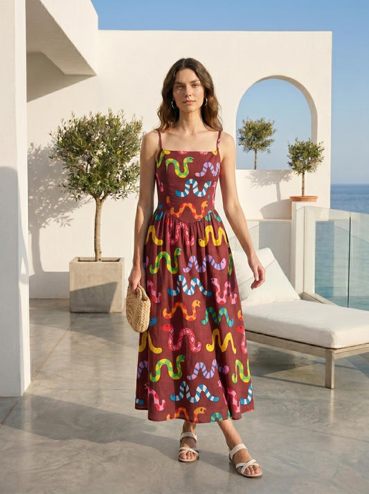 May include: A maroon midi dress with a colourful snake pattern. The dress has thin straps and a square neckline. The woman is holding a small woven bag and wearing white sandals. The background includes a white wall, trees, and a view of the ocean.