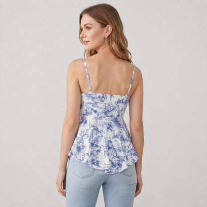 May include: A sleeveless peplum top featuring a white base with a blue toile de jouy floral pattern. The garment has thin shoulder straps, a straight neckline, and a flared hemline that sits at the waist, paired with light blue denim jeans.