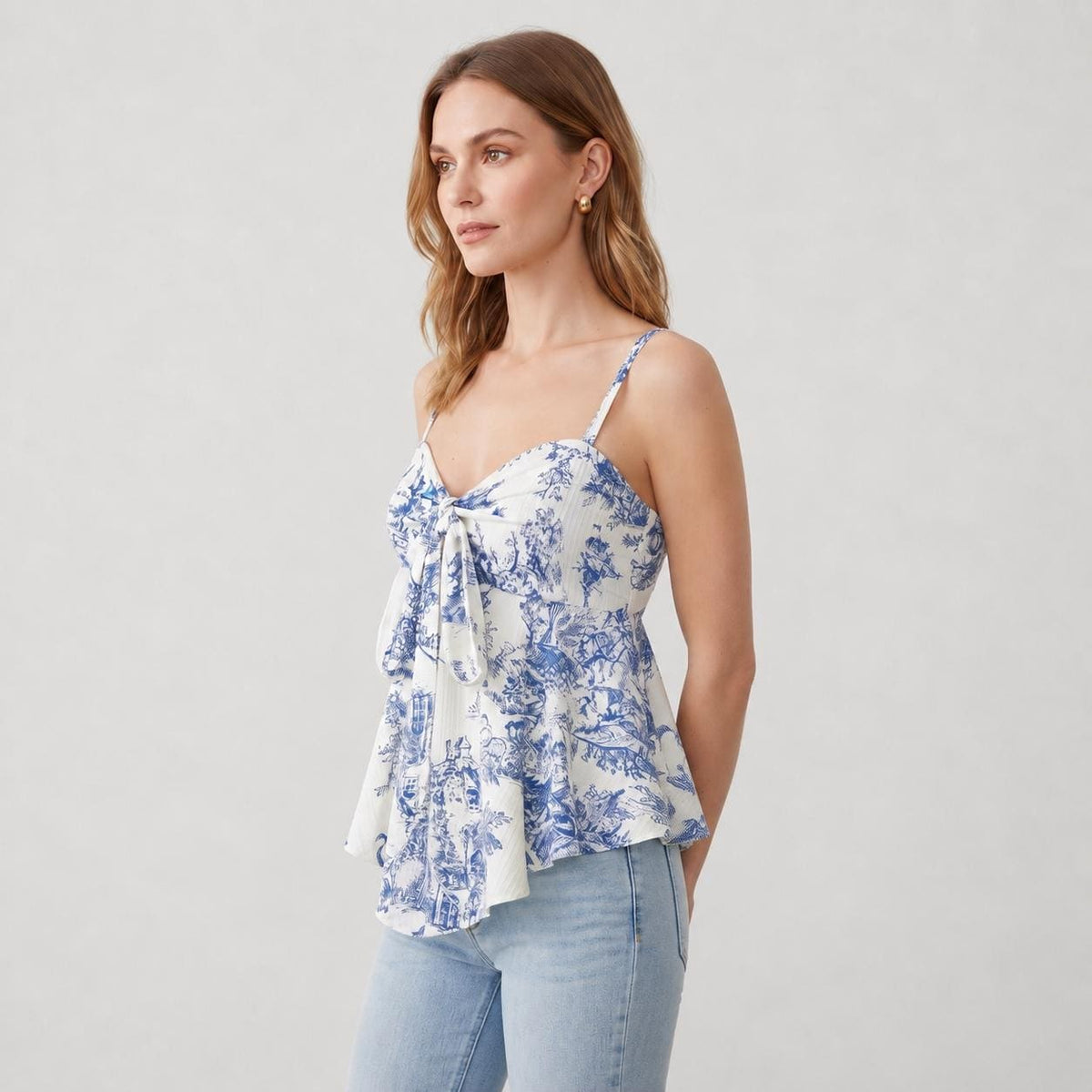 May include: A sleeveless camisole top featuring a white base with a blue toile de jouy pattern. The garment has thin shoulder straps, a sweetheart neckline, and a front tie detail at the bust, creating a flared silhouette that falls at the waist.