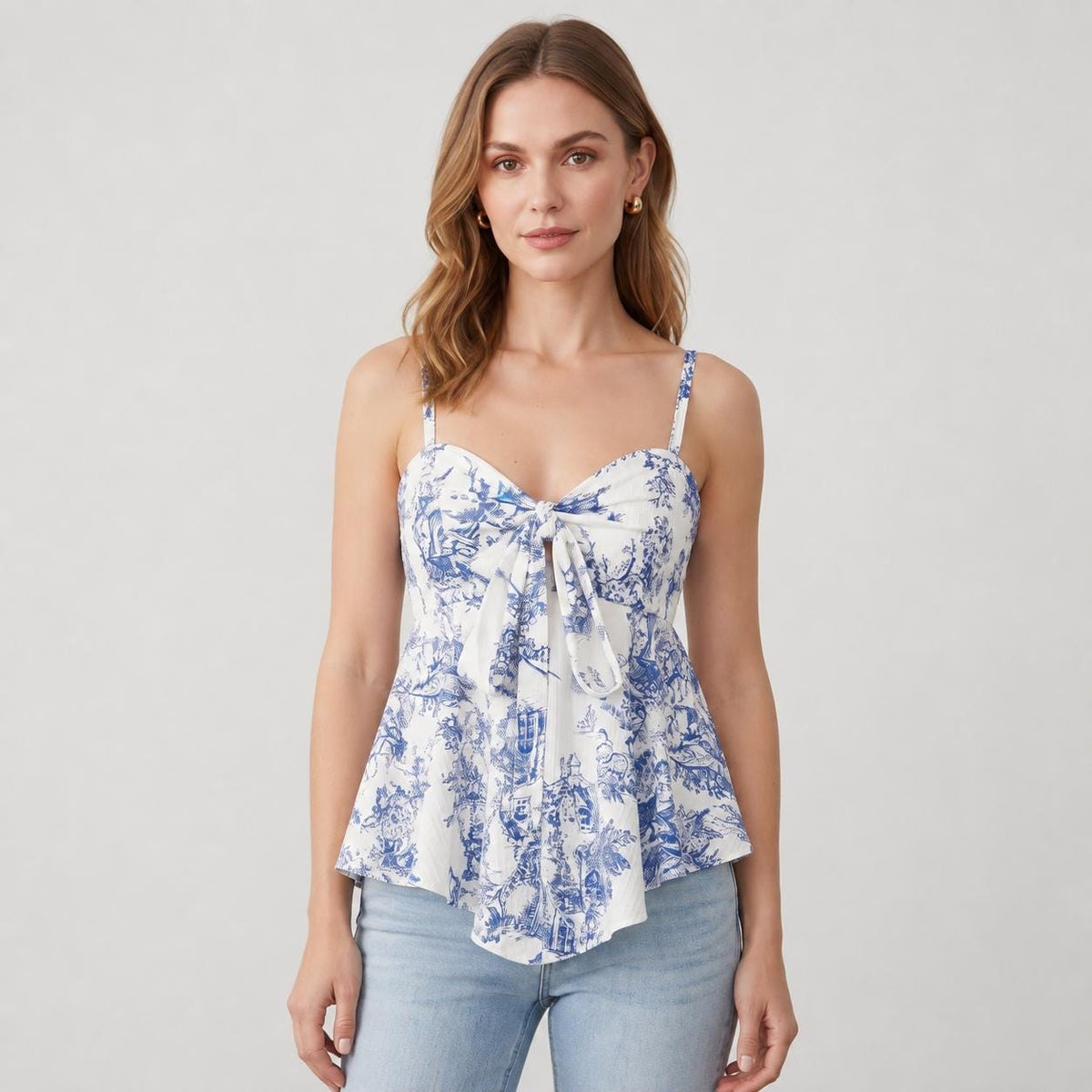 May include: A sleeveless white top featuring a blue toile de jouy pattern, a sweetheart neckline, thin shoulder straps, and a front tie detail that creates a small cutout. The garment has a peplum-style hem and is paired with light blue jeans.