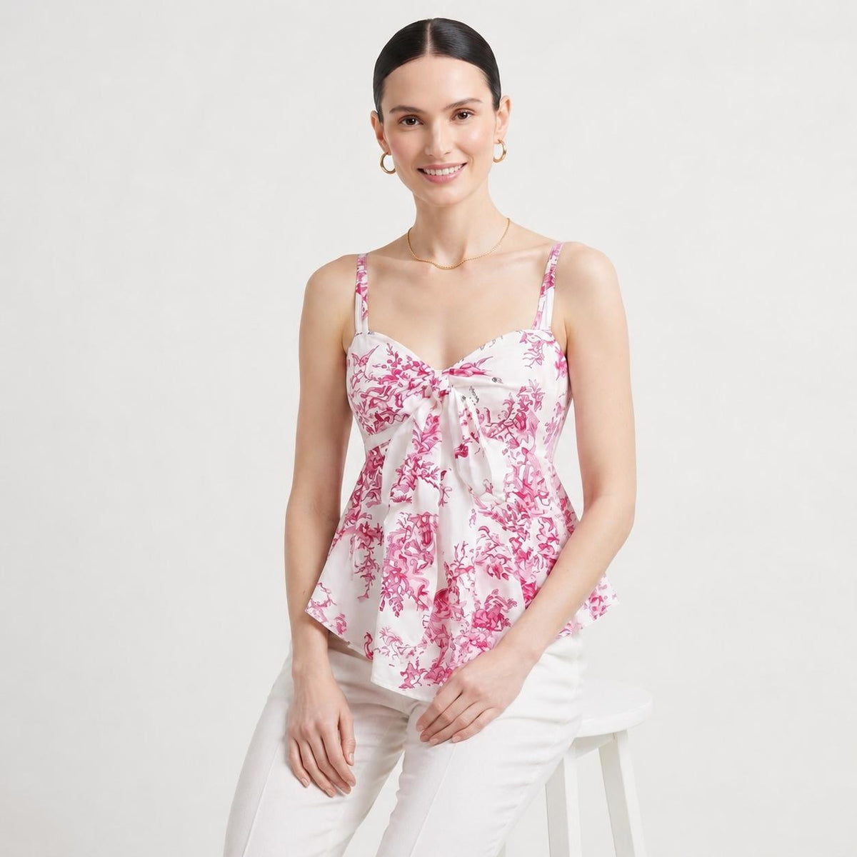 May include: A sleeveless white camisole top featuring a vibrant pink floral toile pattern, a sweetheart neckline, and a central knotted tie detail at the bust. The garment has thin shoulder straps and a slightly flared peplum-style hemline.