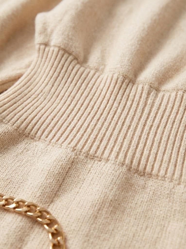 May include: A beige knit sweater featuring a wide, ribbed waistband and a textured fabric surface. A polished gold-tone metal chain link detail is visible at the bottom left corner, resting against the soft, light-colored knit material.