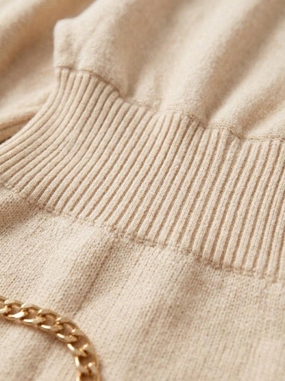 May include: A beige knit sweater featuring a wide, ribbed waistband and a textured fabric surface. A polished gold-tone metal chain link detail is visible at the bottom left corner, resting against the soft, light-colored knit material.
